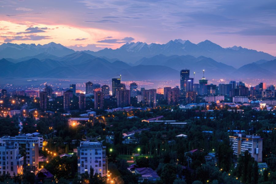 Almaty's cityscape at dusk, with vibrant lights illuminating the buildings and mountains beyond.